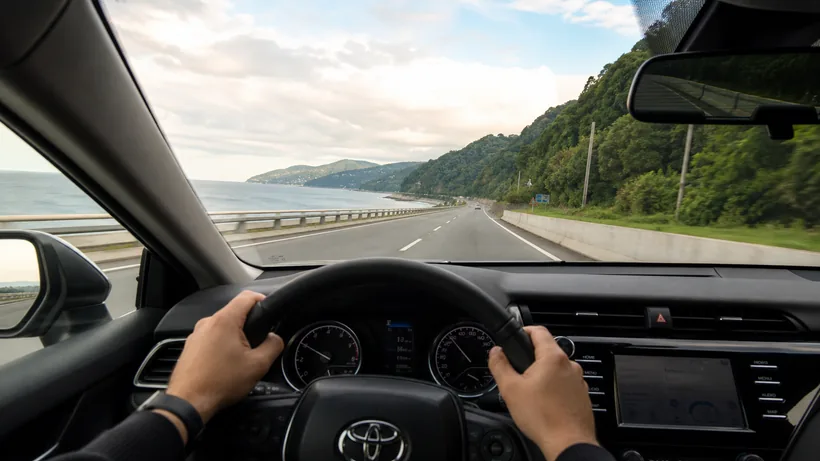A view from the driver seat looking out over a coastal highway in Adjara, with the Black Sea on one side and green hills on the other.